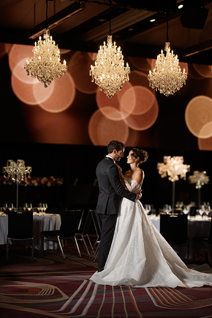 A bride and groom stand together on a decorated ballroom floor under sparkling chandeliers, gazing at each other lovingly during their wedding reception—one of the most cherished event types. Tables are set and the background is softly lit.