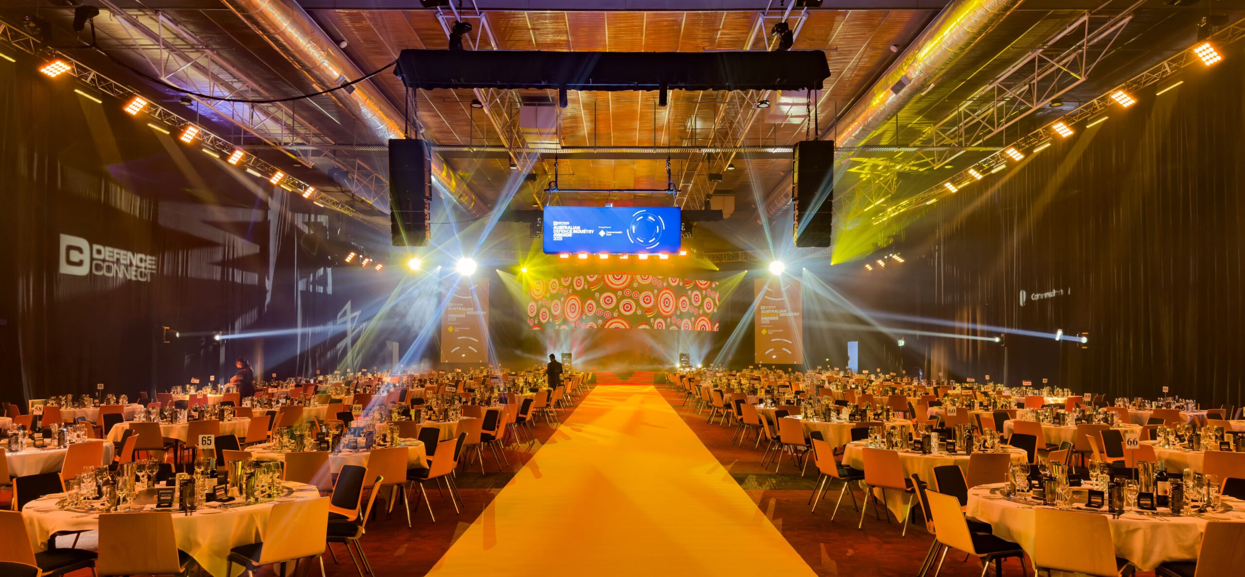 Large event hall with round tables set for dinner, a yellow carpet leading to a brightly lit stage with colorful circular patterns, and spotlights illuminating the room. Screens and logos are visible on the walls.