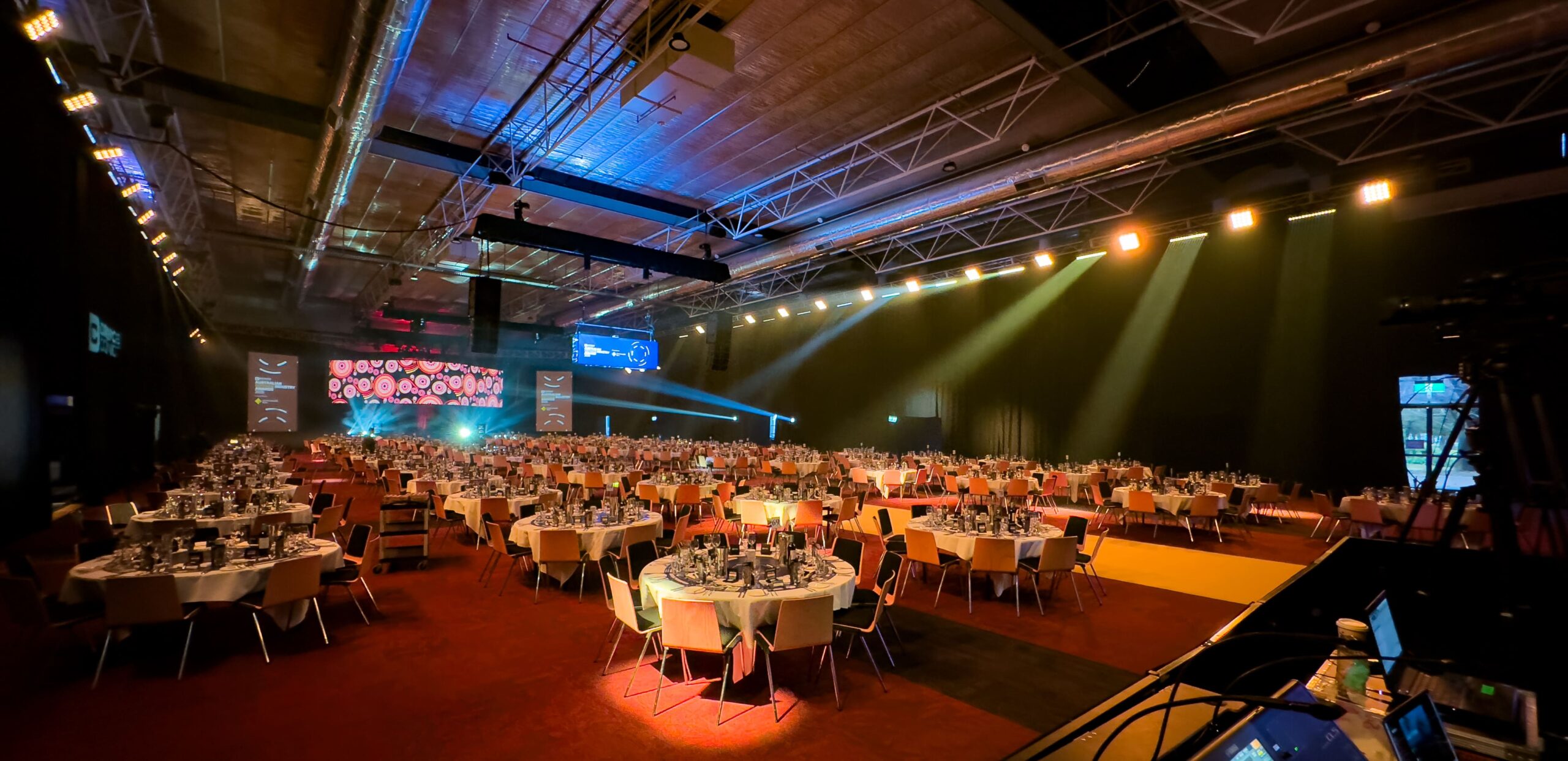 A large banquet hall with round tables set for an event, each table covered with white tablecloths and elegant place settings. Colorful stage lighting and video screens illuminate the spacious, modern venue.