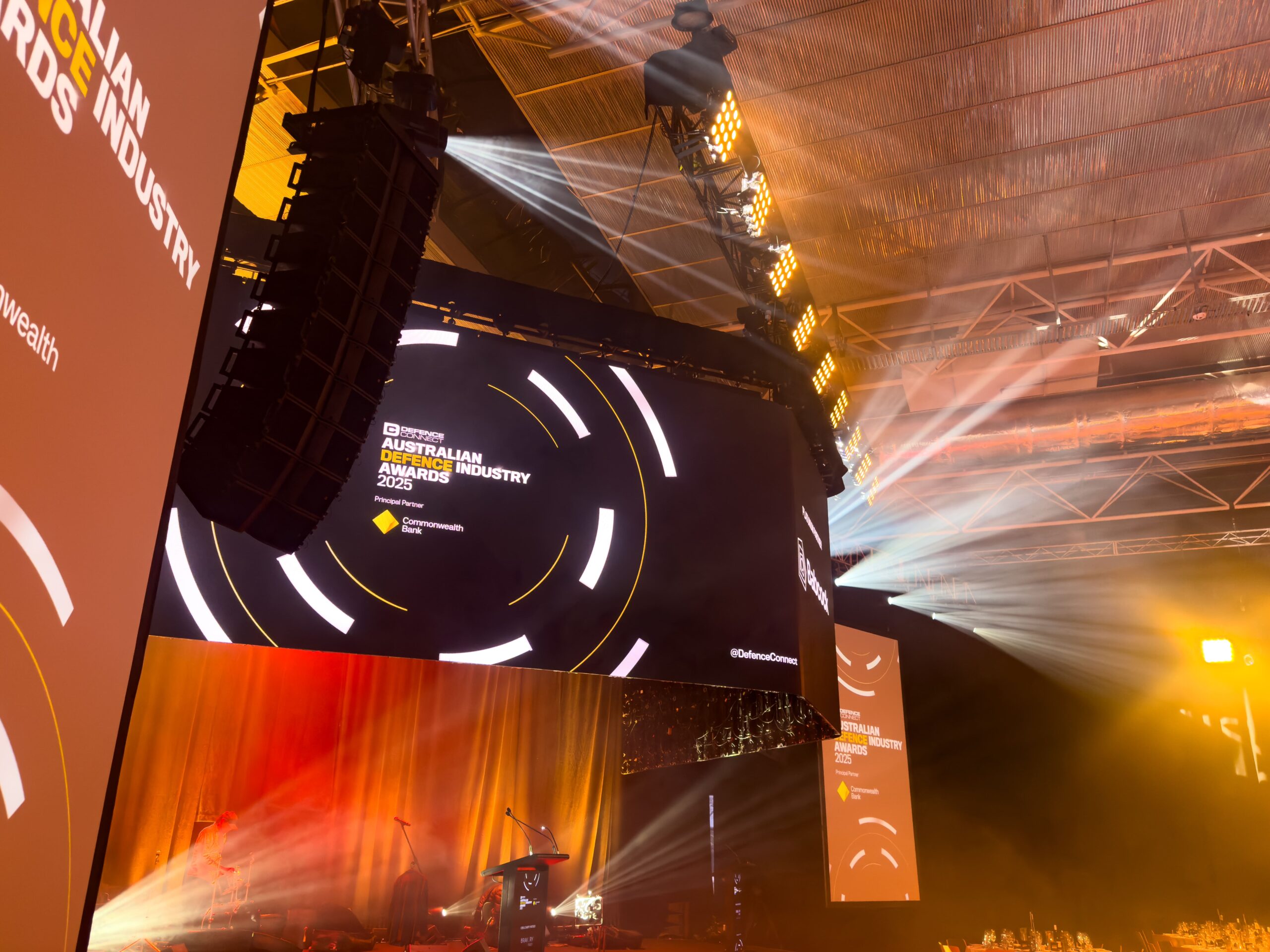 A large screen at an indoor event displays Australian Defence Industry Awards 2025 with stage lights, sound equipment, and spotlights illuminating the space. The venue ceiling and part of the audience are visible.