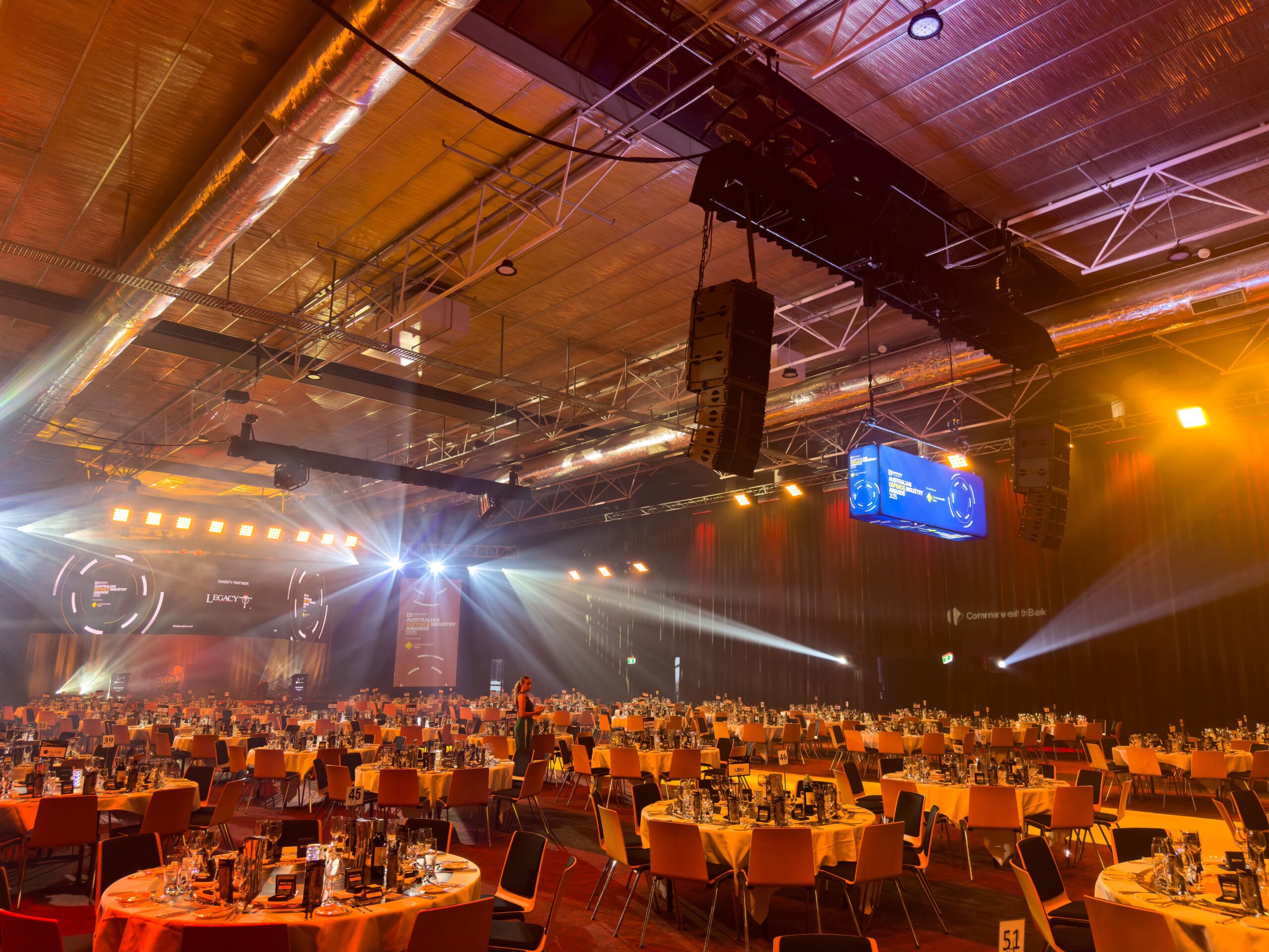 A large event hall with round tables covered in orange tablecloths, set for a formal dinner. Stage lights and screens are lit, and a few people are walking among the tables. The ceiling has exposed pipes and speakers.