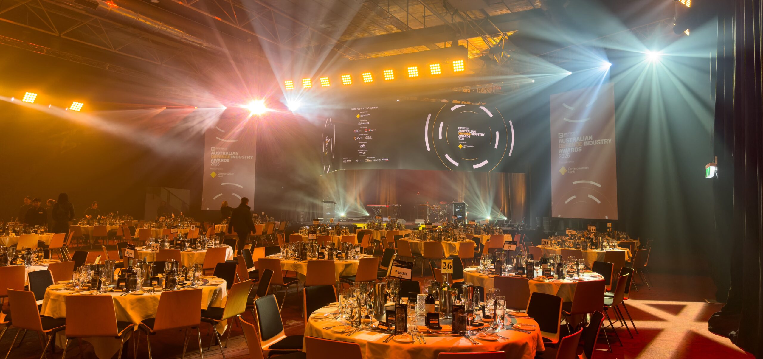 A large, elegant event hall set up for a gala dinner, with round tables covered in yellow tablecloths, glassware neatly arranged, stage lighting, and screens displaying event graphics in the background.