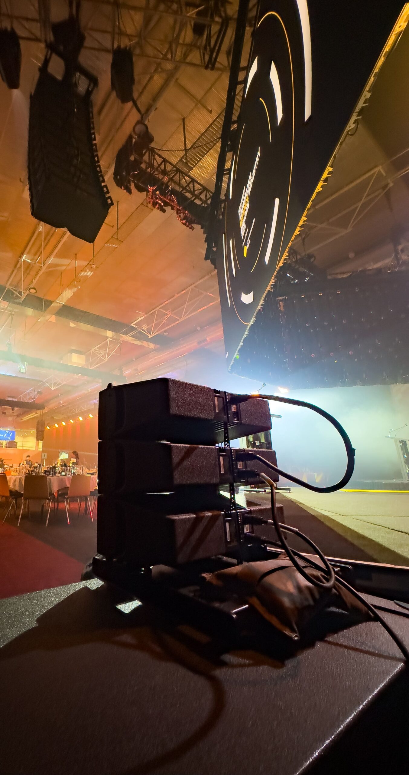 Stacked electronic equipment with connected cables sits on a table in the foreground of a dimly lit event space, with a large screen, stage lights, and seating area visible in the background.
