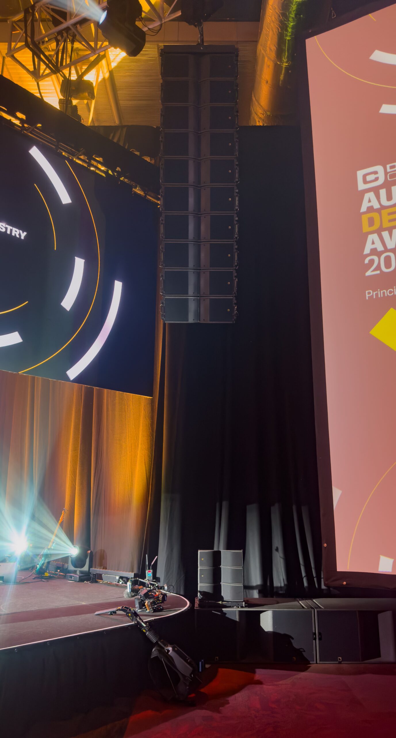 A stage setup with orange and yellow lighting, large speaker stacks, and a screen displaying geometric designs. Some stage equipment is visible near the front.