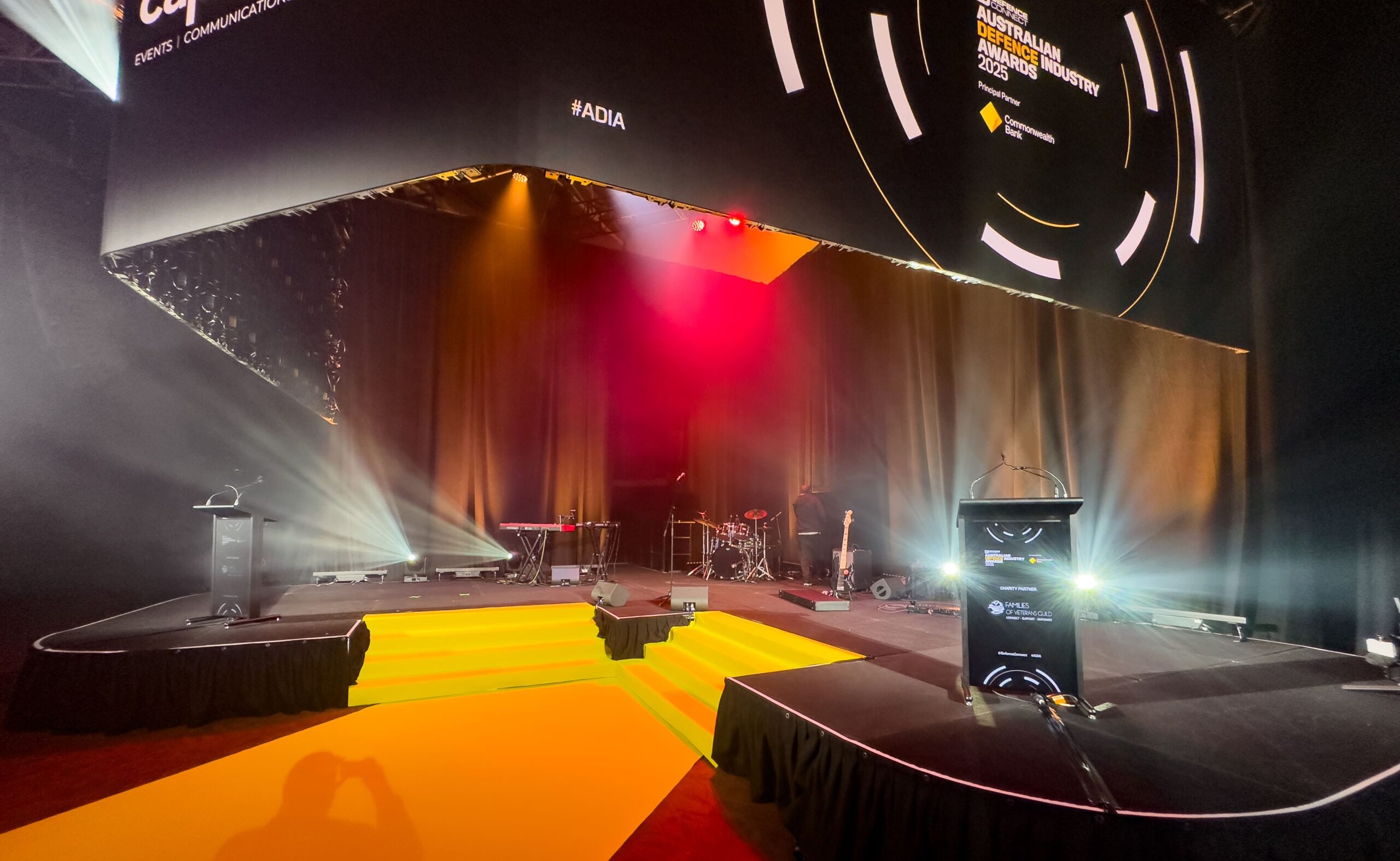A stage with yellow steps, two podiums, and musical instruments under colorful lighting at the Australian Defence Industry Awards event. A large circular sign with event branding hangs above the stage.