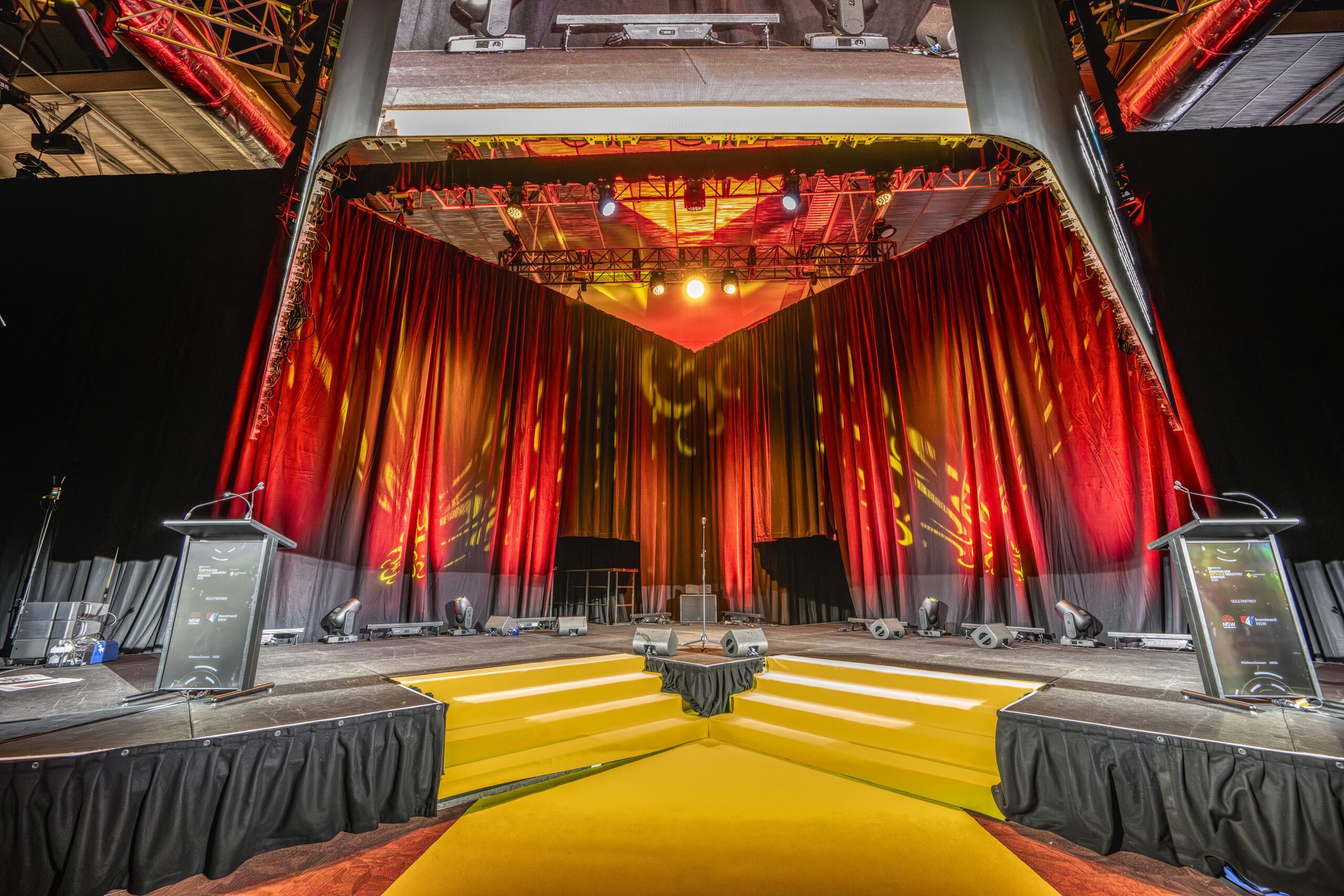 A brightly lit stage with red curtains, yellow spotlights, and two podiums on either side. The stage is elevated with stairs in the center, and lighting equipment is visible on the floor and above.