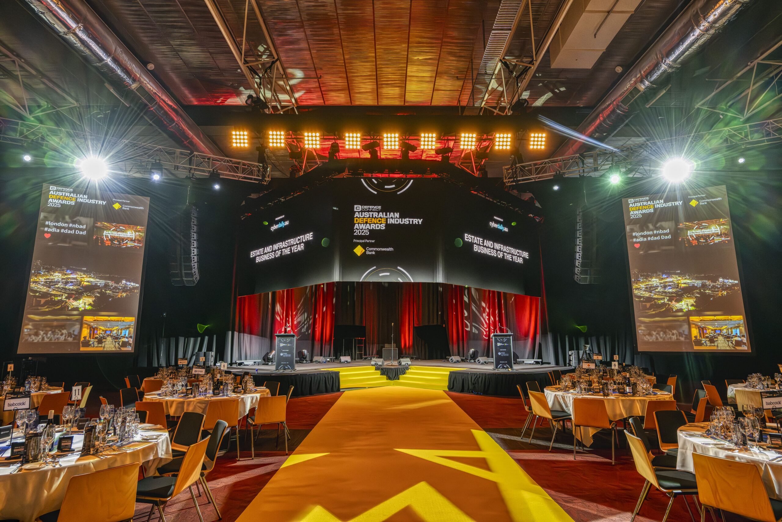 A large, decorated banquet hall set up for an awards ceremony, with round dining tables on both sides, a bright yellow carpet leading to a stage, and big screens displaying event details on the walls.