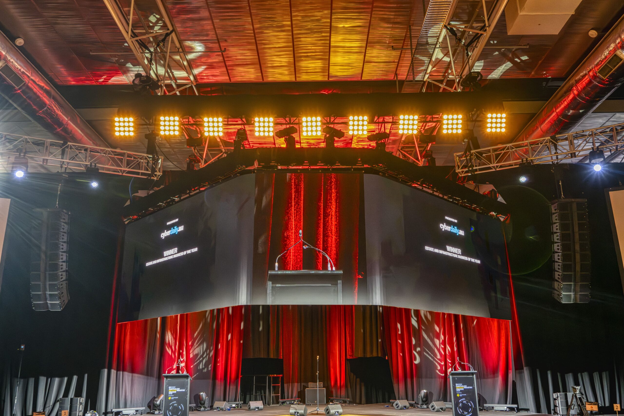 A large stage with red and black curtains, two big screens, a podium at the center, and bright stage lights illuminating the area, set for an event or presentation in a spacious indoor venue.