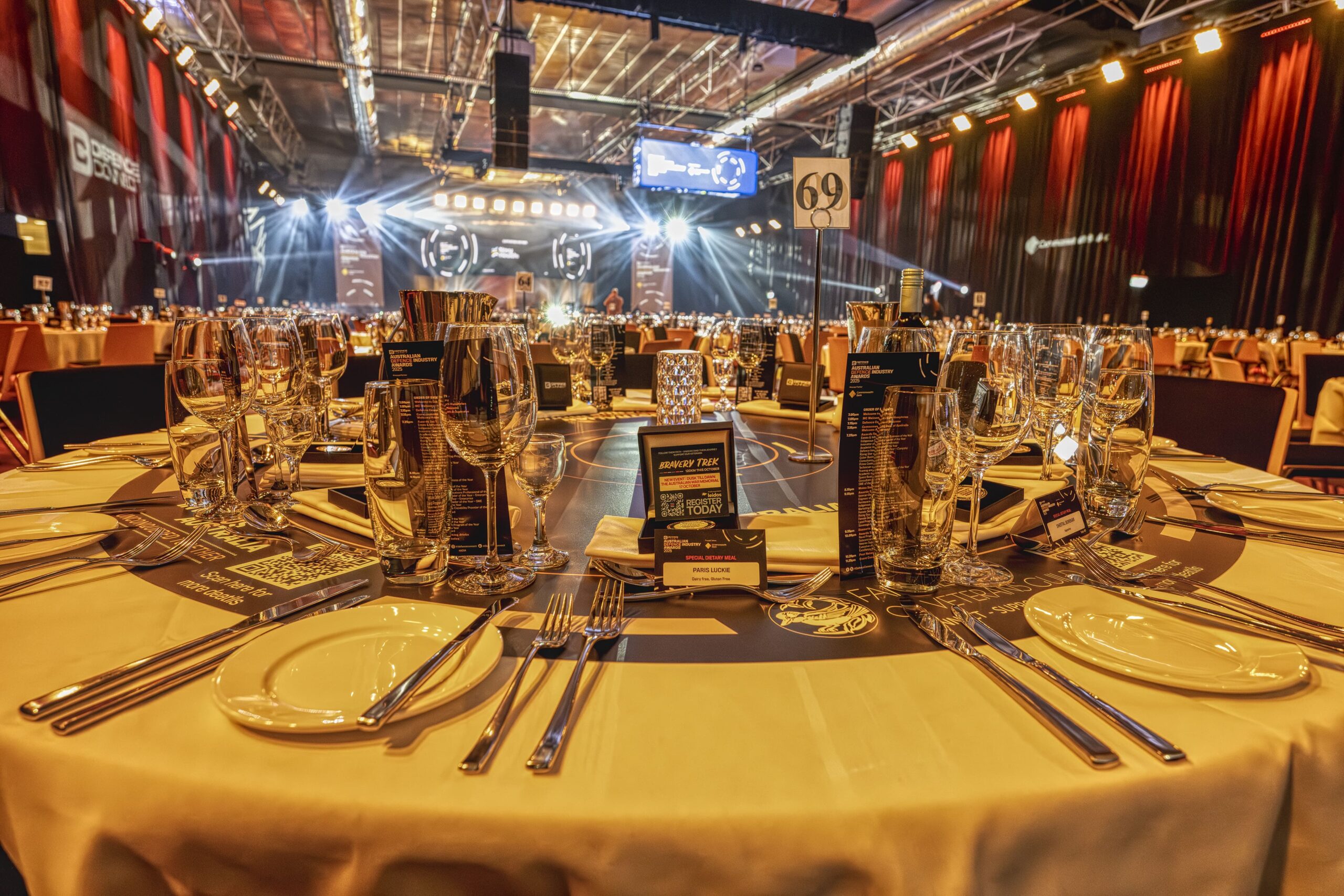 A round banquet table set with glassware, plates, cutlery, and menus in a large, elegant event hall with stage lights, hanging drapes, and the table number 69 visible. Other tables and the stage appear in the background.