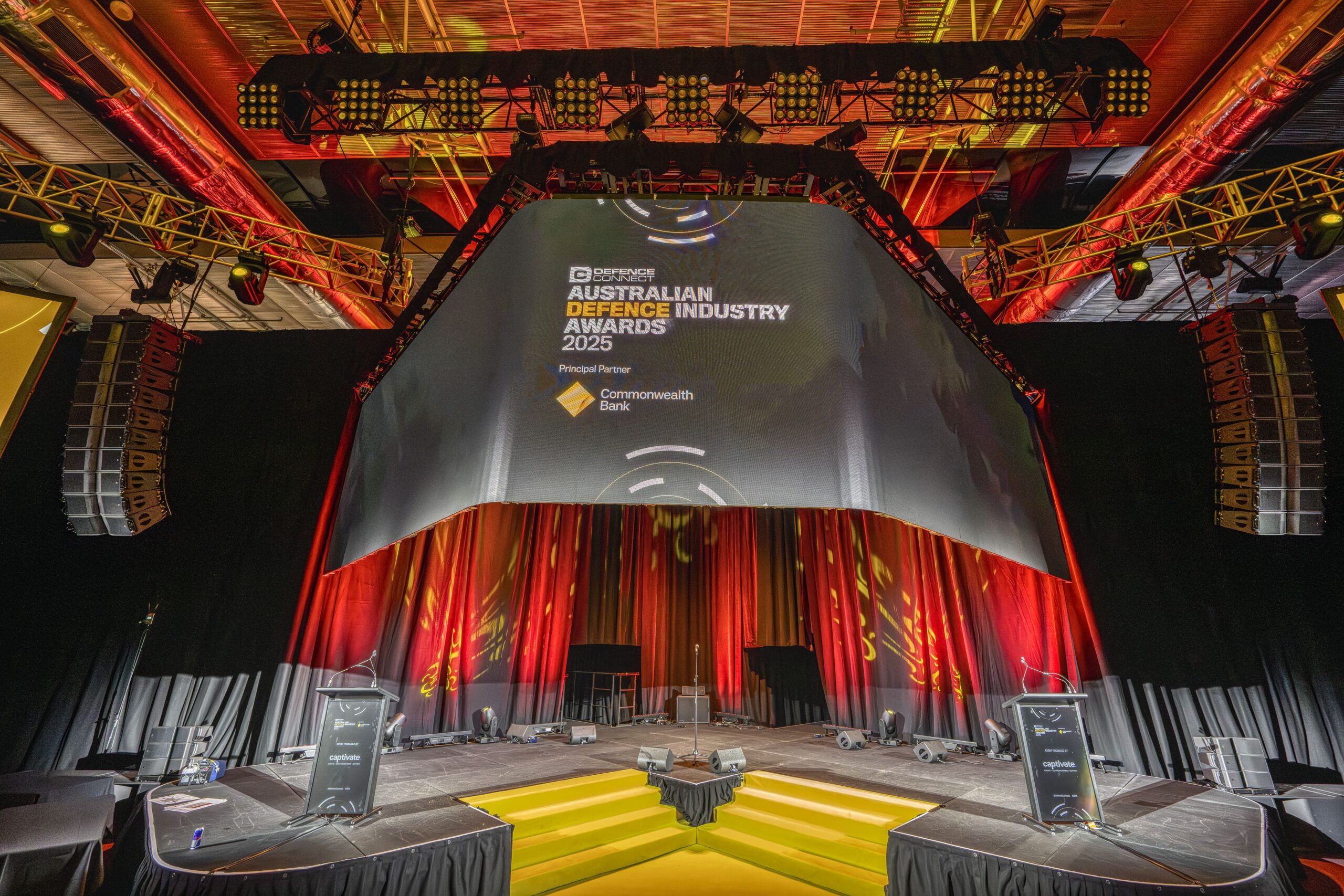 A stage setup for the Australian Defence Industry Awards 2025, featuring a large screen displaying the event name and sponsor, with dramatic lighting, speakers, podiums, and red and yellow accents.