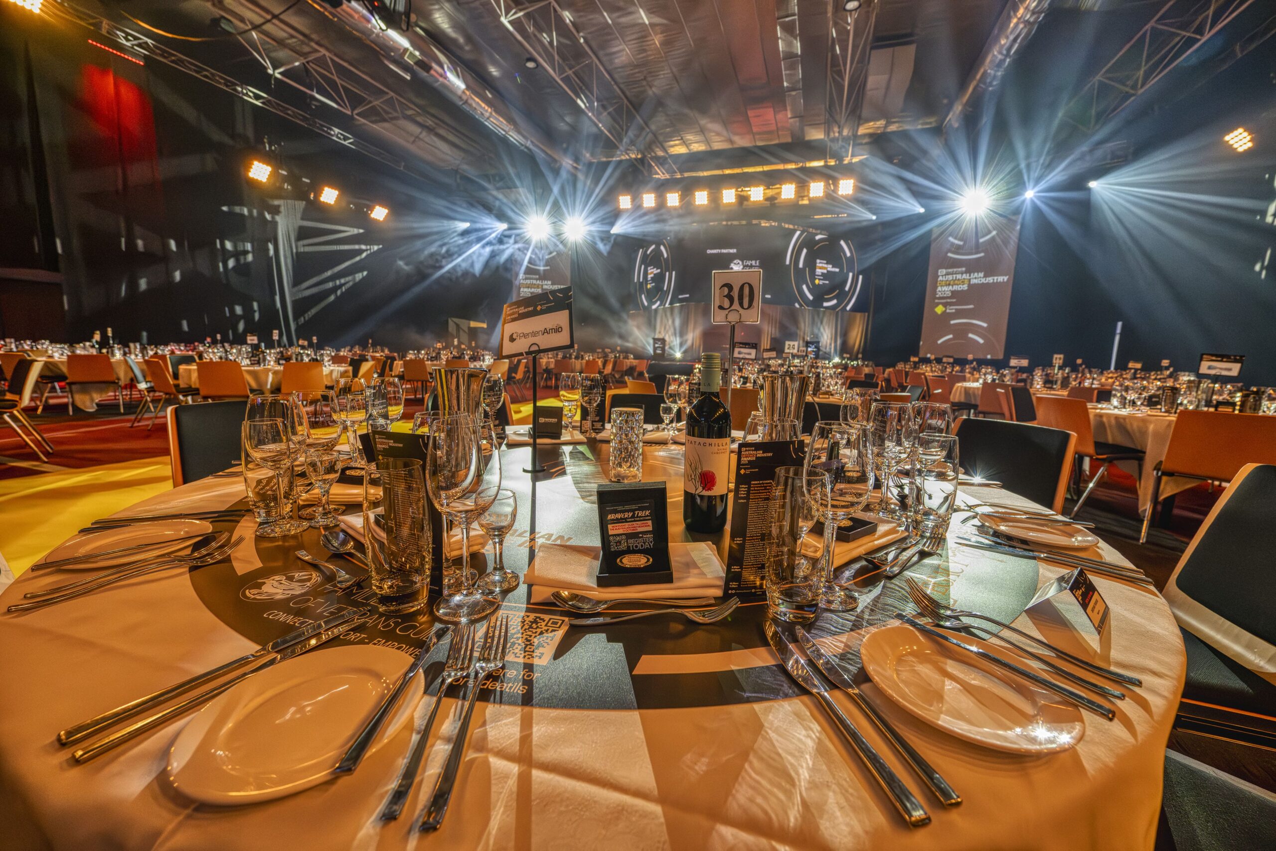 A round banquet table set with glassware, plates, and silverware is in the foreground of a large, elegant event hall with bright stage lights and many similarly set tables in the background.