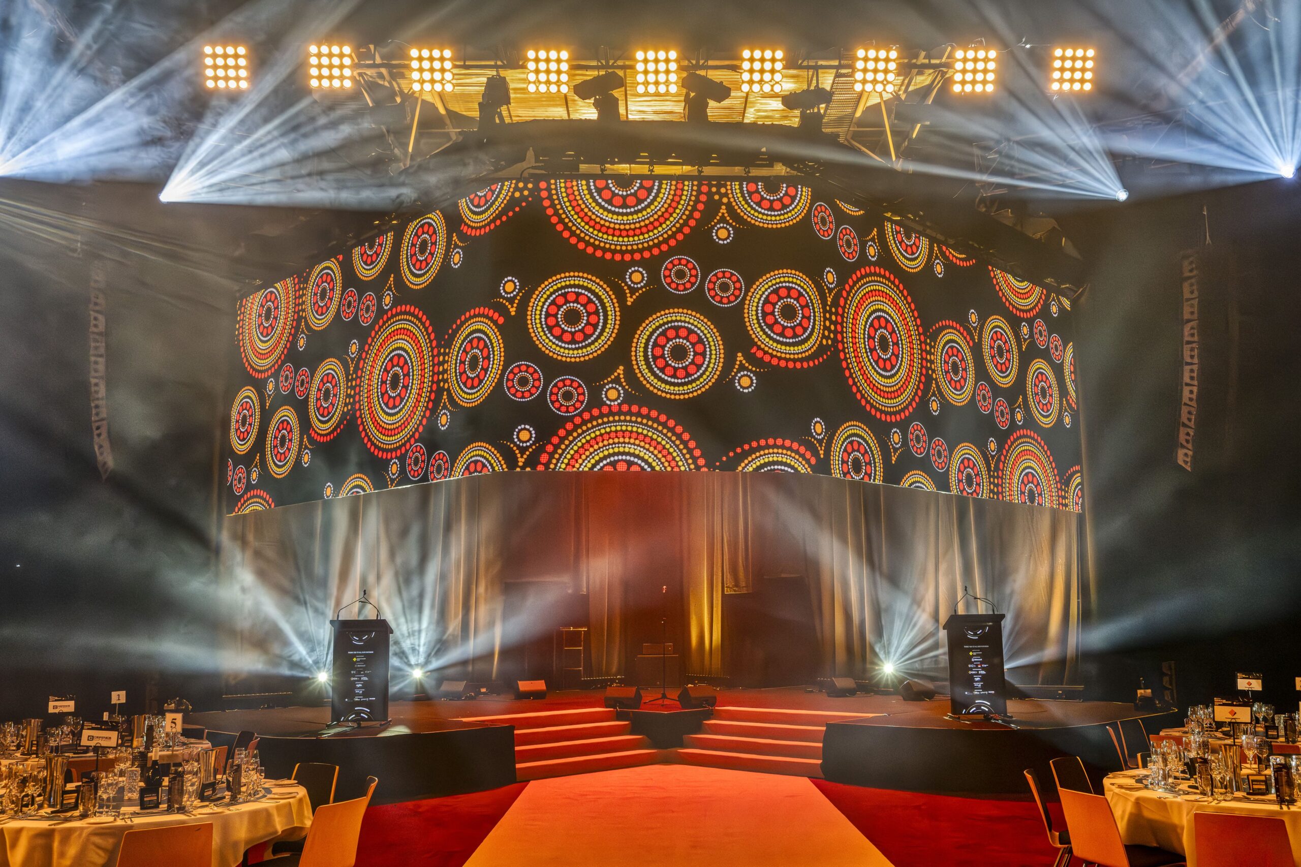 A brightly lit stage with red carpeted stairs, decorated tables in the foreground, and a large digital screen displaying colorful, circular Aboriginal-inspired patterns. Spotlights shine down, creating a dramatic atmosphere.
