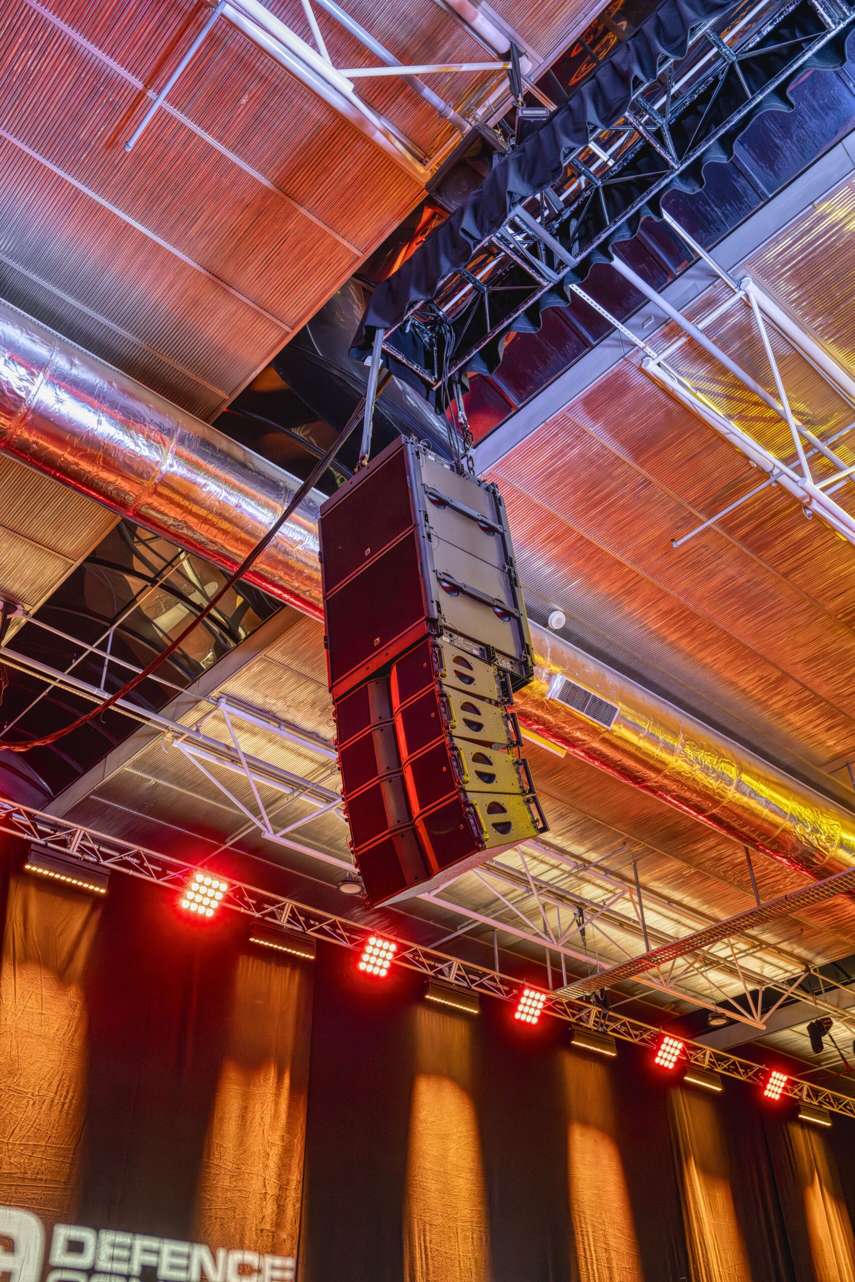 A line array of black speakers is suspended from the ceiling in an indoor event venue with exposed metal beams, ductwork, and red stage lights illuminating the scene.