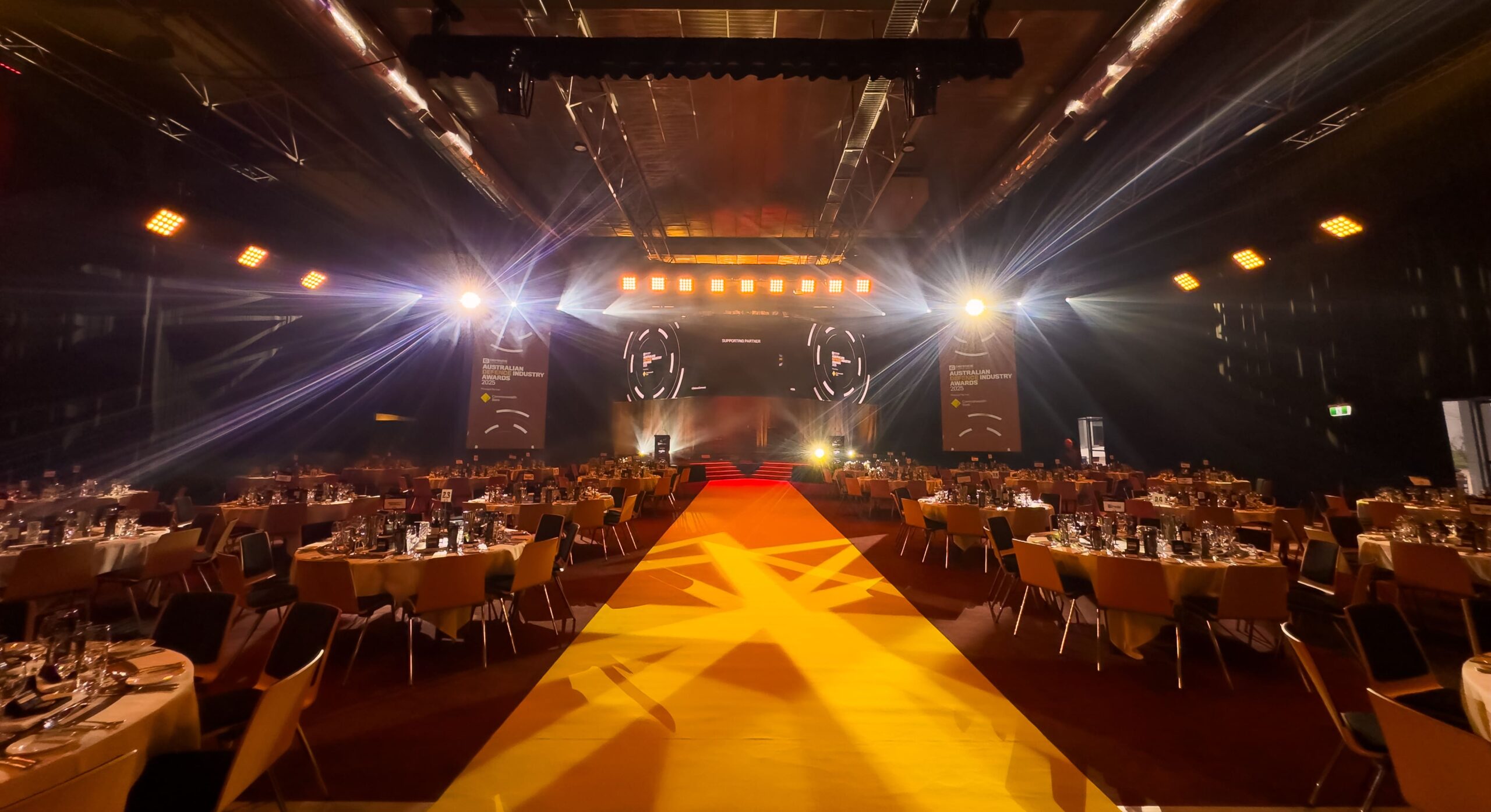 A large, elegant event hall set up for a formal dinner, with round tables covered in white cloths, stage with bright spotlights, and a yellow carpet leading to the stage at the front.