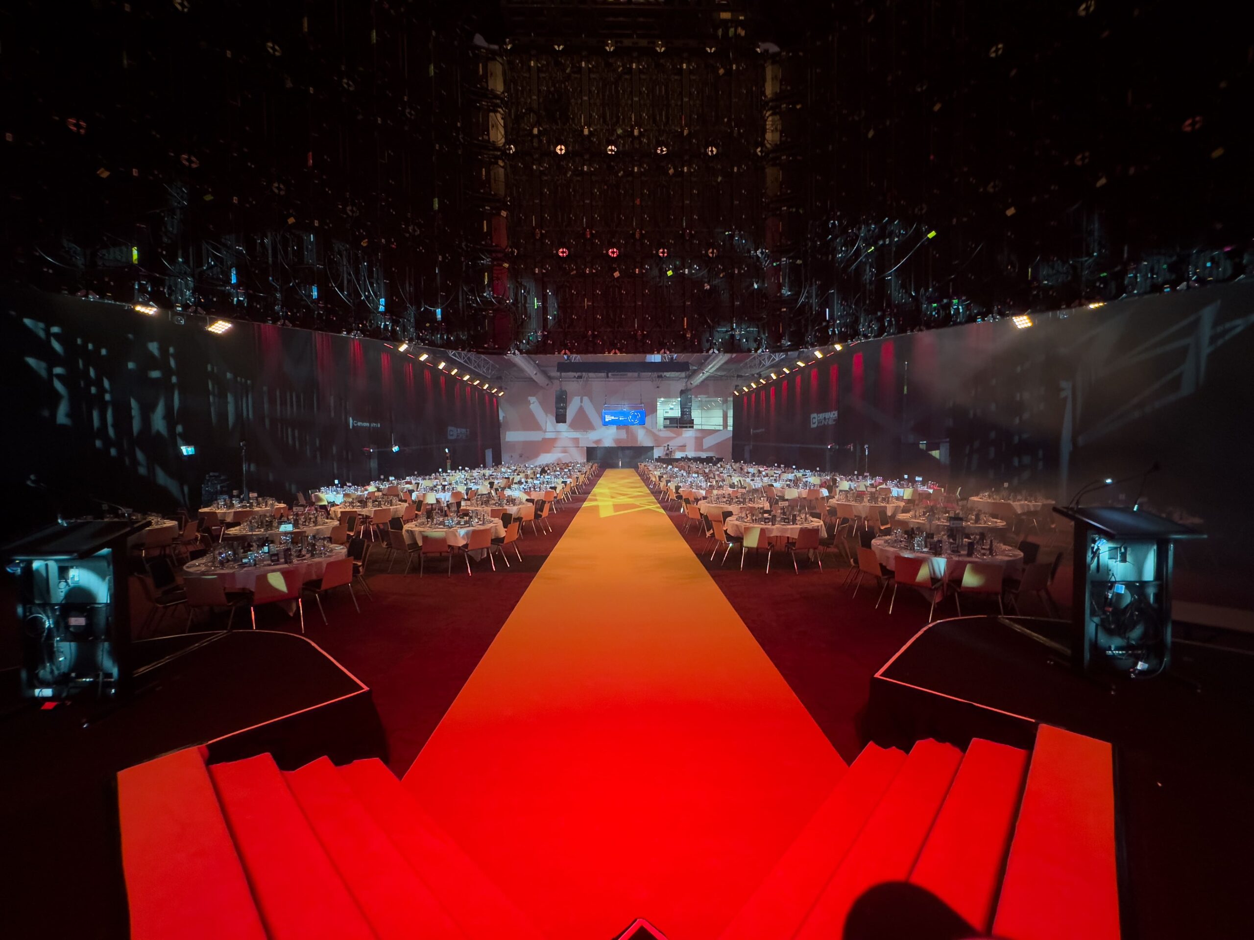 A wide-angle view of an elegant banquet hall with round tables set for an event, a bright yellow carpet in the center aisle, dramatic lighting, and large screens at the far end of the room.
