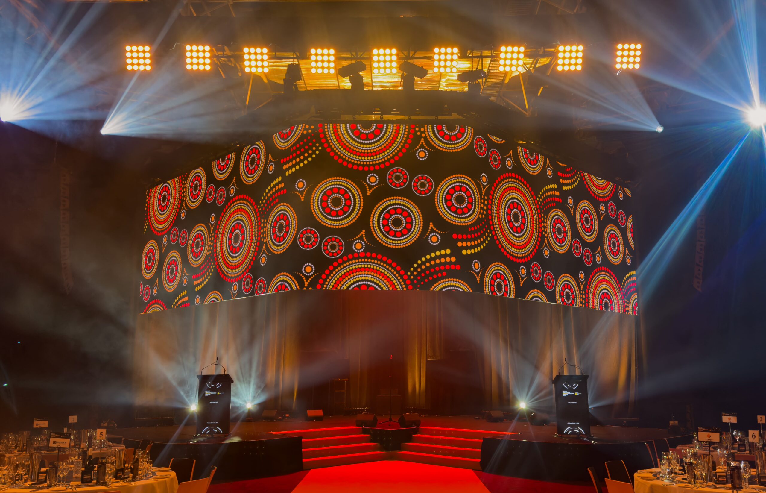 A stage with red carpeted steps and a large, curved screen displaying vibrant Aboriginal-inspired circular dot patterns. Bright lights shine from above, illuminating the scene. Tables set for an event are visible in the foreground.