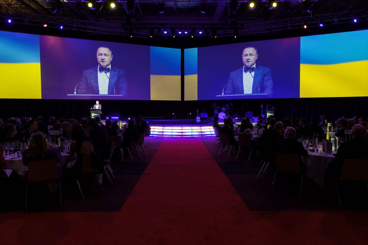 A man in a tuxedo speaks at a podium on a stage at ADIA 2023, with large screens behind him displaying his image and the Ukrainian flag; an audience sits at tables in a dimly lit banquet hall.