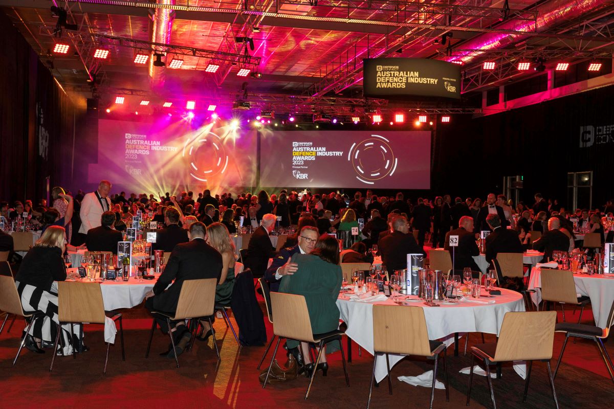 A large crowd sits at round tables in a banquet hall decorated with red lights, attending the ADIA 2023 Australian Defence Industry Awards event. Two large screens display the event name at the front of the room.