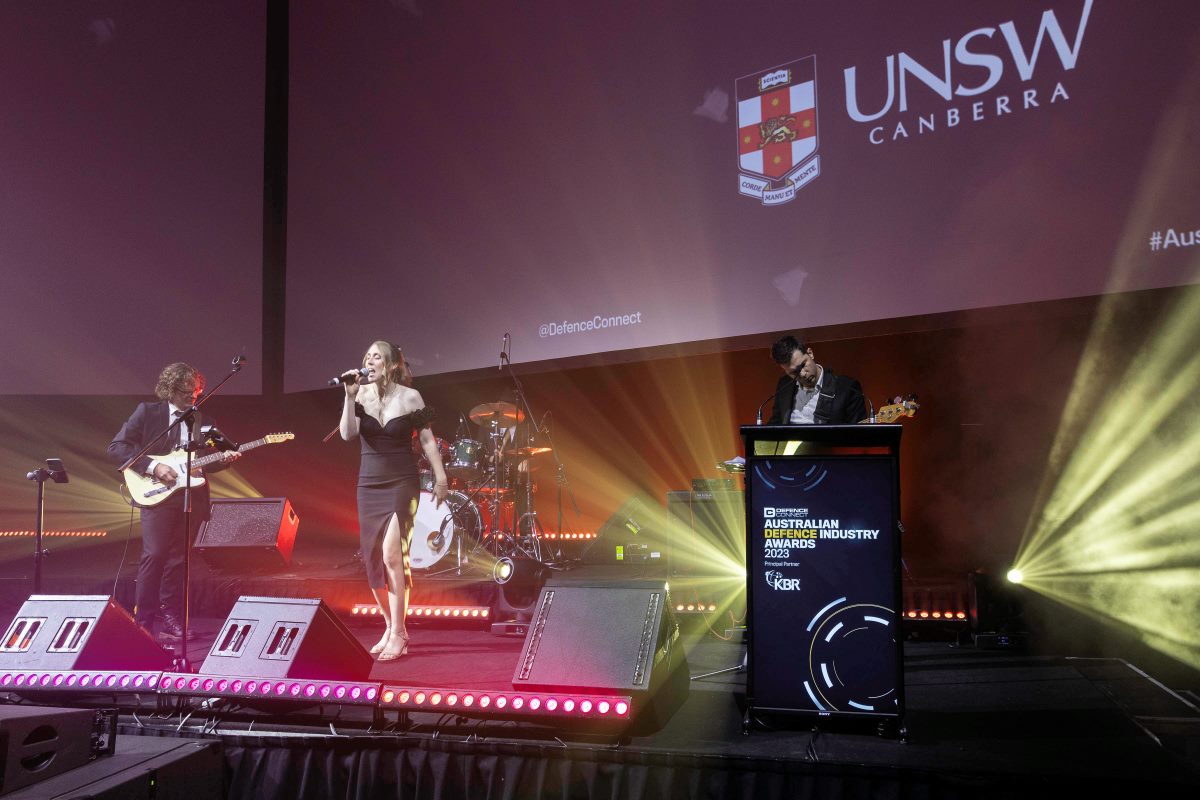 A live band performs on stage at ADIA 2023, featuring a female singer, guitarist, drummer, and keyboardist. The backdrop displays the UNSW Canberra logo and event information, with colorful stage lighting illuminating the scene.