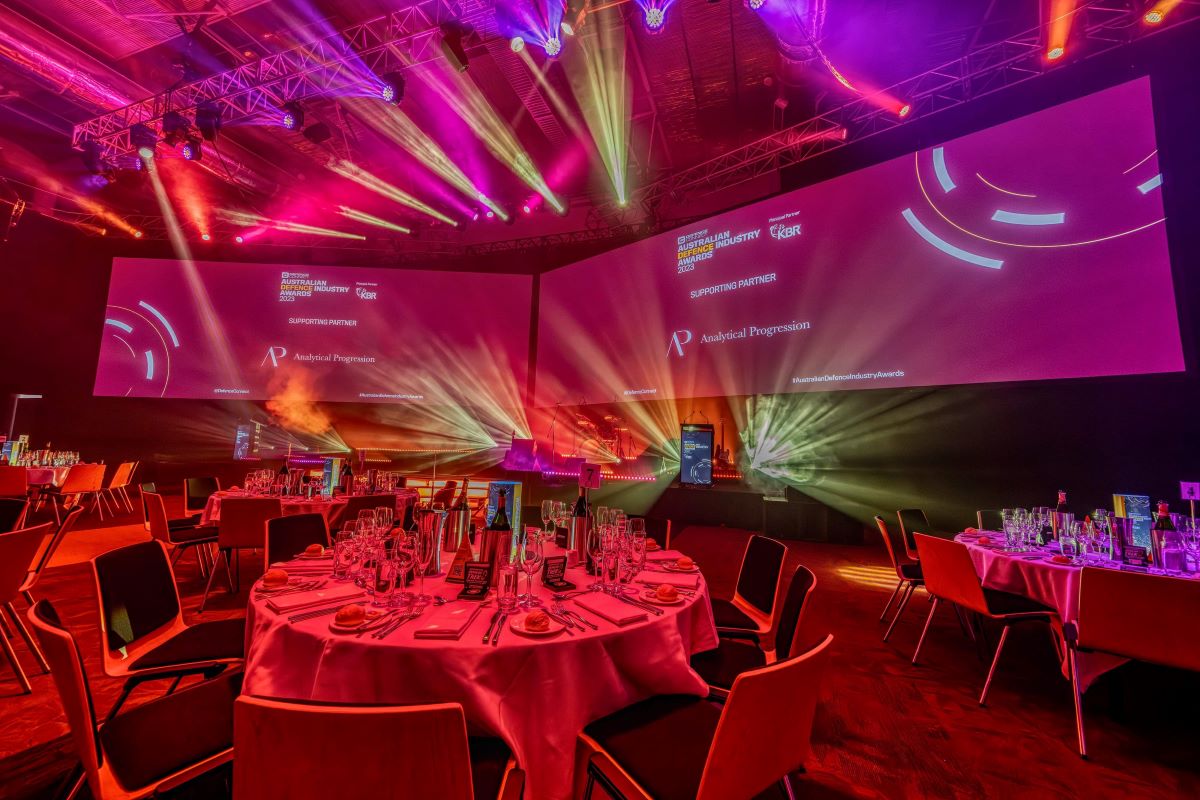 A vibrant banquet hall with round tables set for dinner, dramatic colorful lighting, and large projection screens displaying ADIA 2023 event graphics and sponsor logos on the walls.