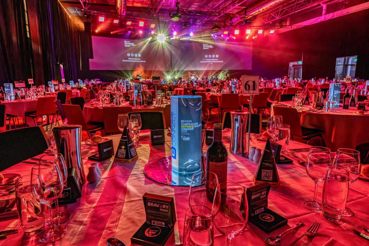 A banquet hall with round tables set for an event, featuring wine bottles, glasses, and menus under red and purple lighting. The ambiance of ADIA 2023 fills the space, with a stage, large screens, and spotlights in the background.