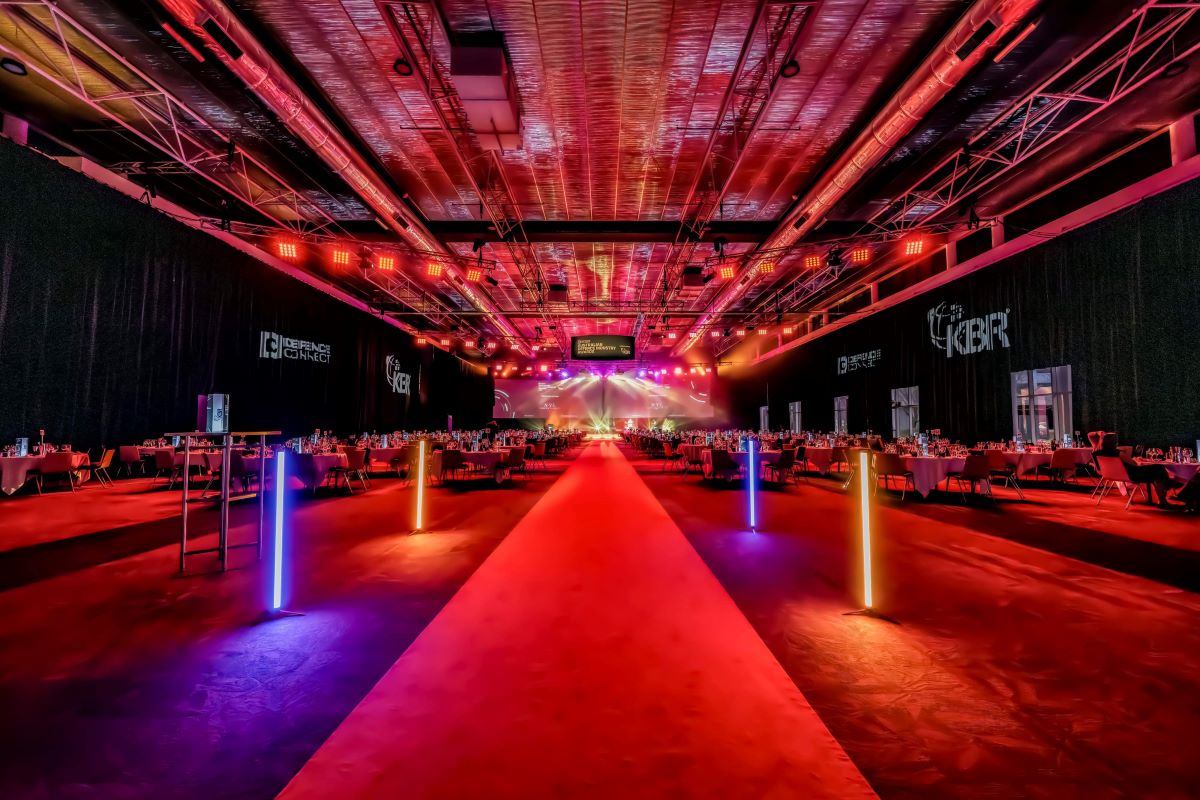A large, modern event hall with a red carpet aisle, neon lights, and dinner tables on both sides sets the scene for ADIA 2023. The stage at the end is brightly lit with colorful spotlights, creating a vibrant, festive atmosphere.