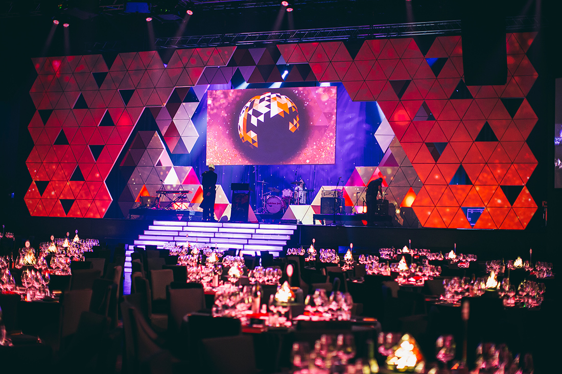 A large, colorful stage set for a gala dinner features geometric red and orange patterns, a big screen displaying a graphic, and tables set with glassware and candles illuminating the dimly lit audience area.