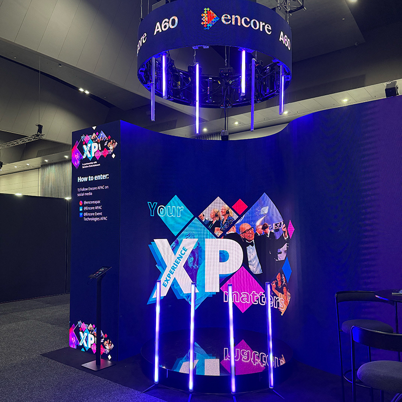 A vibrant event booth features a large screen with “Your XP matters” surrounded by colorful graphics, under a circular sign reading “encore.” Instructions for entering a contest are displayed on a digital board.