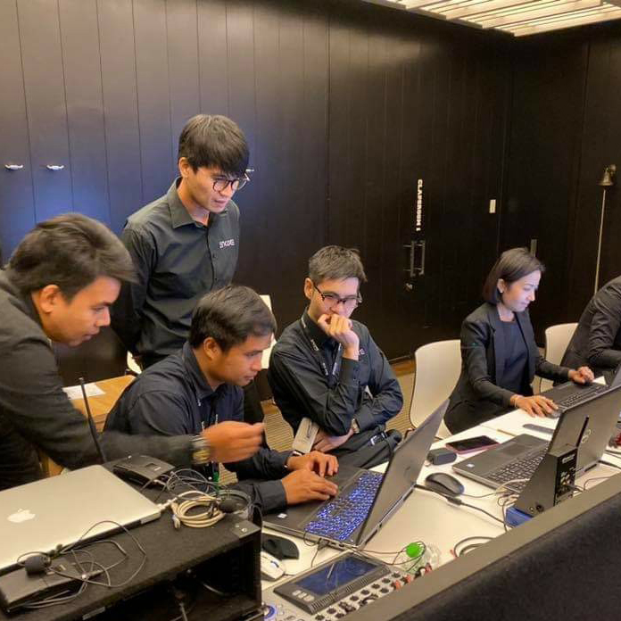 Five people in black attire work together at a long table with laptops and electronics, appearing focused and engaged in discussion—capturing the behind-the-scenes energy of an event production company in Thailand.