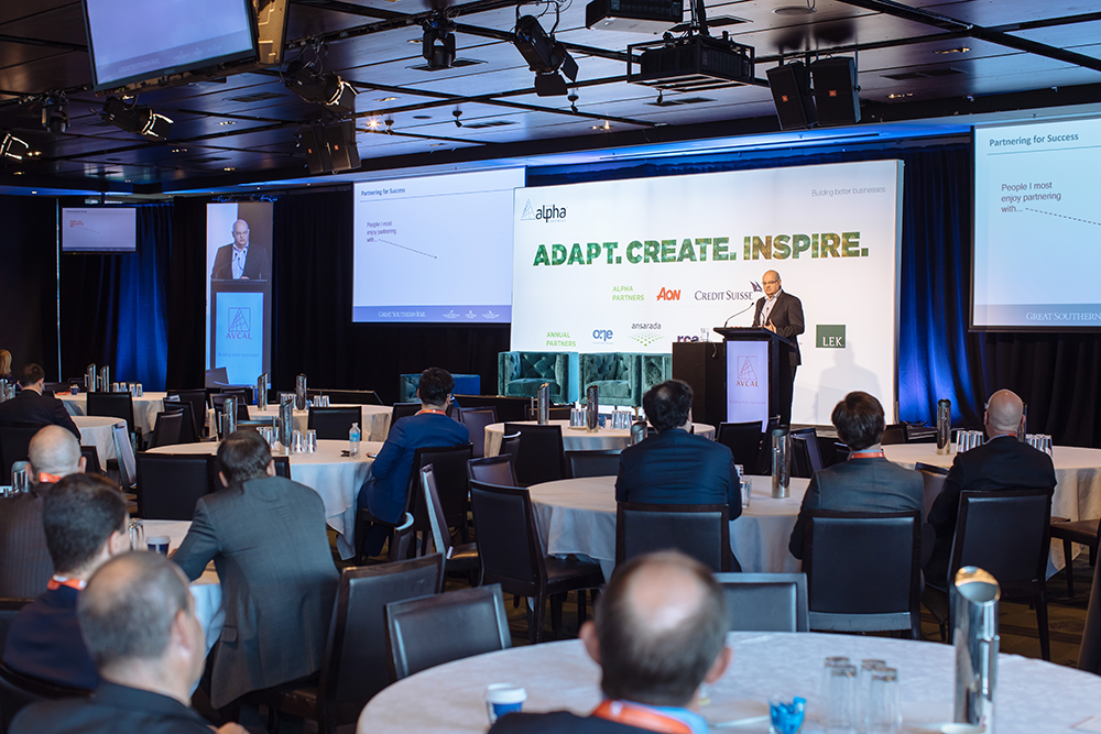A speaker stands at a podium addressing an audience in a modern conference room with round tables, hosting a conference and gala dinner. Behind him, a large screen displays ADAPT. CREATE. INSPIRE. and a slide about partnering for success.
