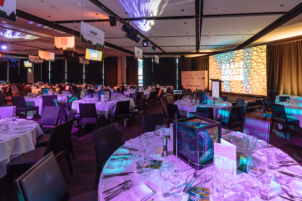 A large, modern banquet hall set for hosting a conference and gala dinner, with round tables covered in white cloths, place settings, and glassware. A brightly lit stage displays the words LEAD CREATE INSPIRE in colorful text.