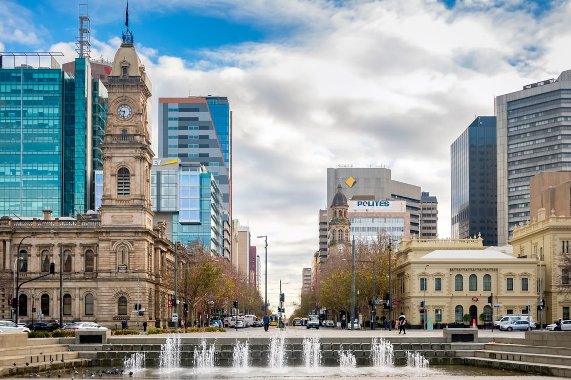 A cityscape featuring historic and modern buildings, a clock tower on the left, and a fountain in the foreground, with cars and trees lining the street under a partly cloudy sky.