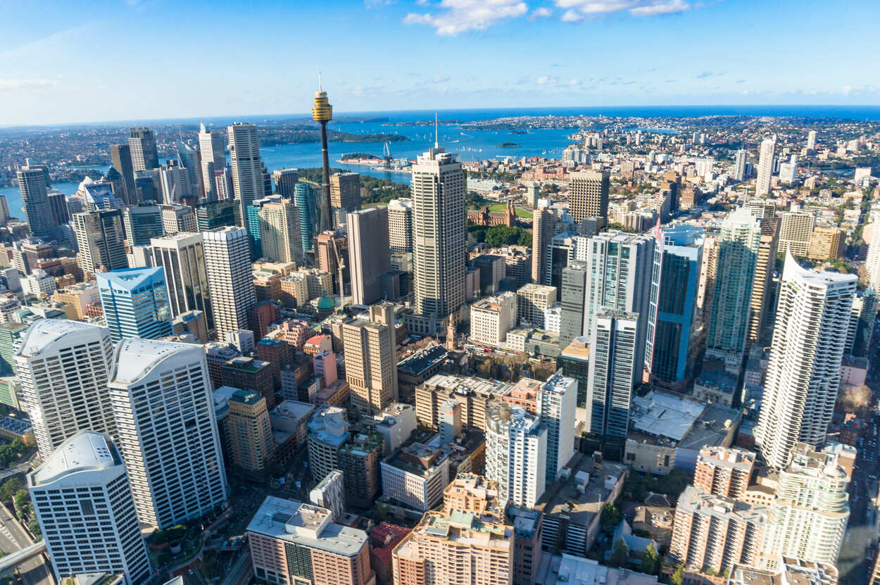 Aerial view of a cityscape with many tall buildings, a harbor with blue water in the background, and clear skies above.