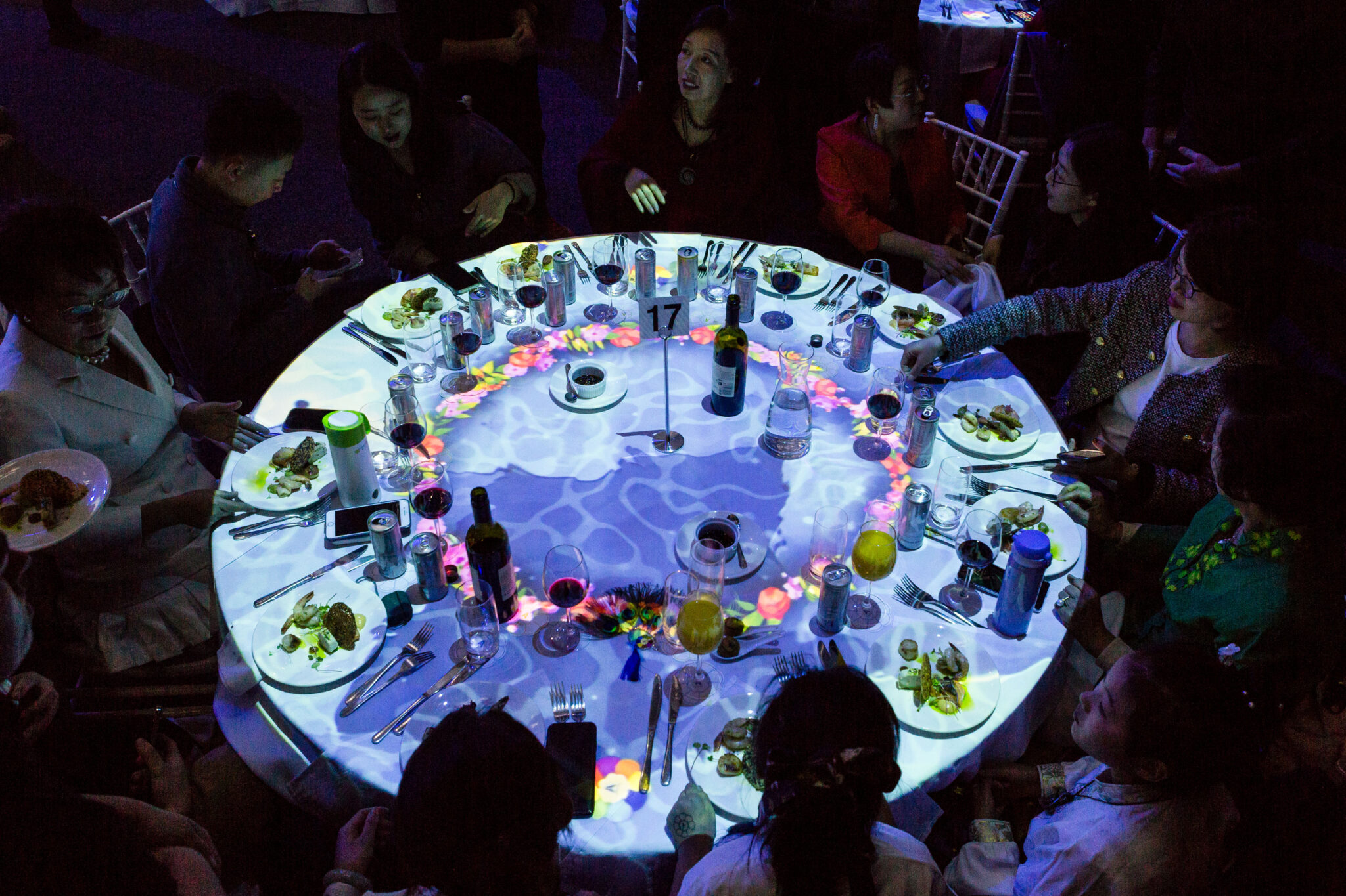 A group of people sit around a round table with plates of food, while colorful lights and patterns are projected onto the table’s surface, creating a vibrant, interactive display.
