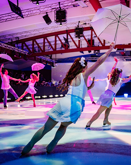 Figure skaters in white costumes perform on an ice rink, each holding a white umbrella. Colorful lights illuminate the scene, creating a vibrant, theatrical atmosphere perfect for an unforgettable encore.