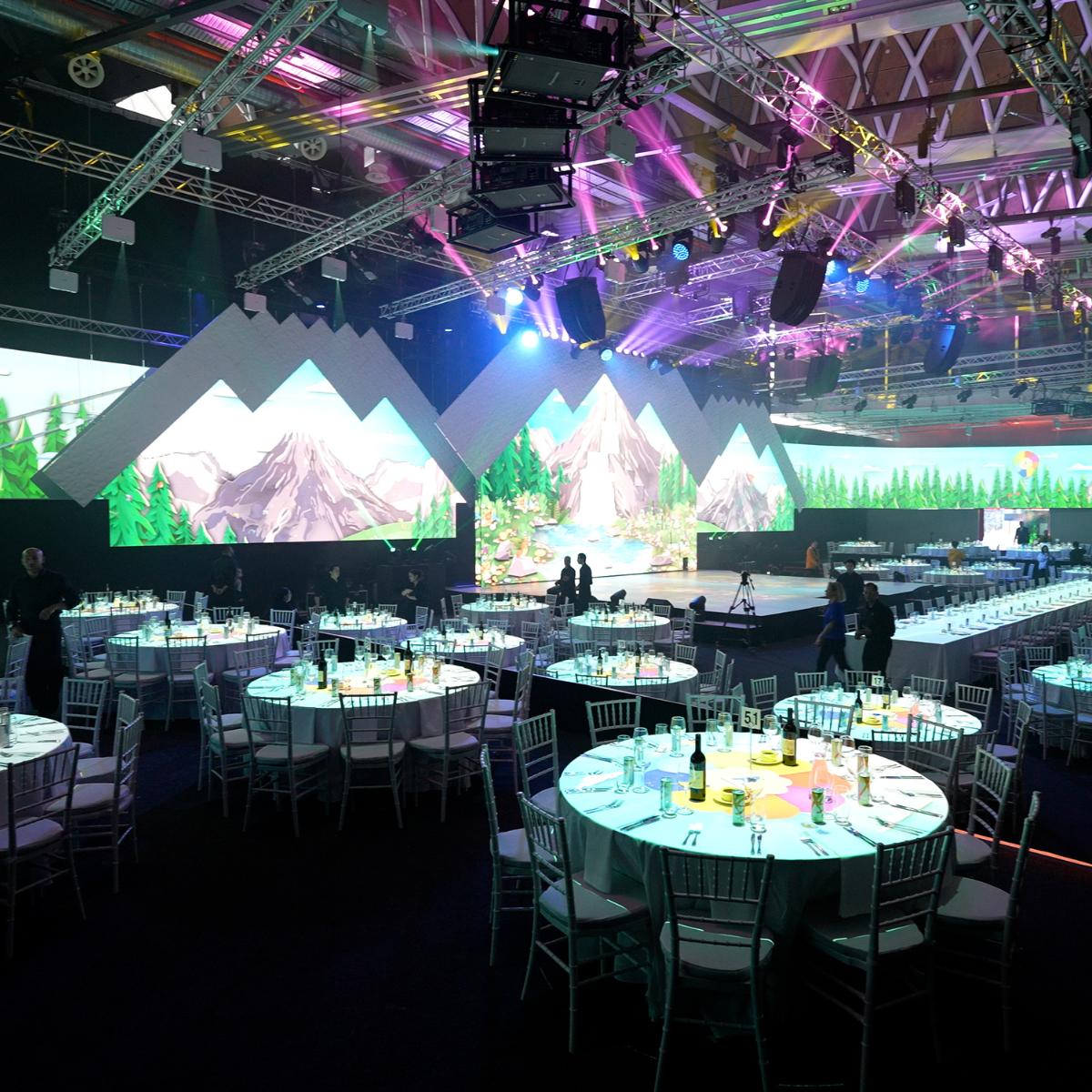 A large event hall set for a formal dinner, with round tables and white chairs. The stage features a backdrop of stylized mountains and trees, and colorful lights illuminate the ceiling and room.