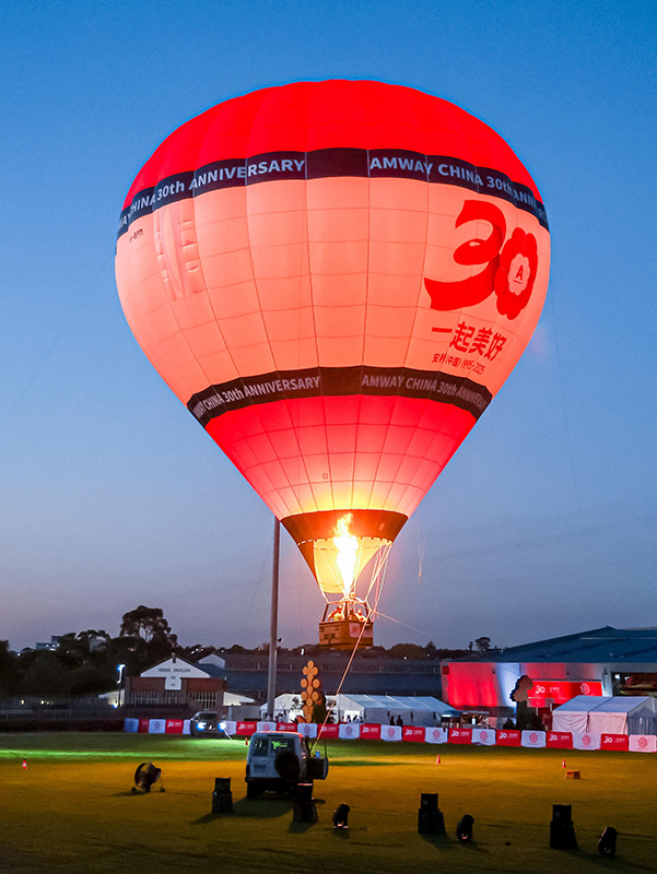 A large red and white hot air balloon with “Amway China 30th Anniversary” and “30” printed on it is illuminated by its burner at dusk, floating above a grassy field near buildings.