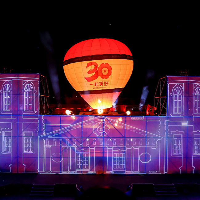 A brightly lit hot air balloon with 30 and Chinese text floats above a stage decorated with colorful lights and outlined drawings of buildings, creating a festive nighttime celebration atmosphere.