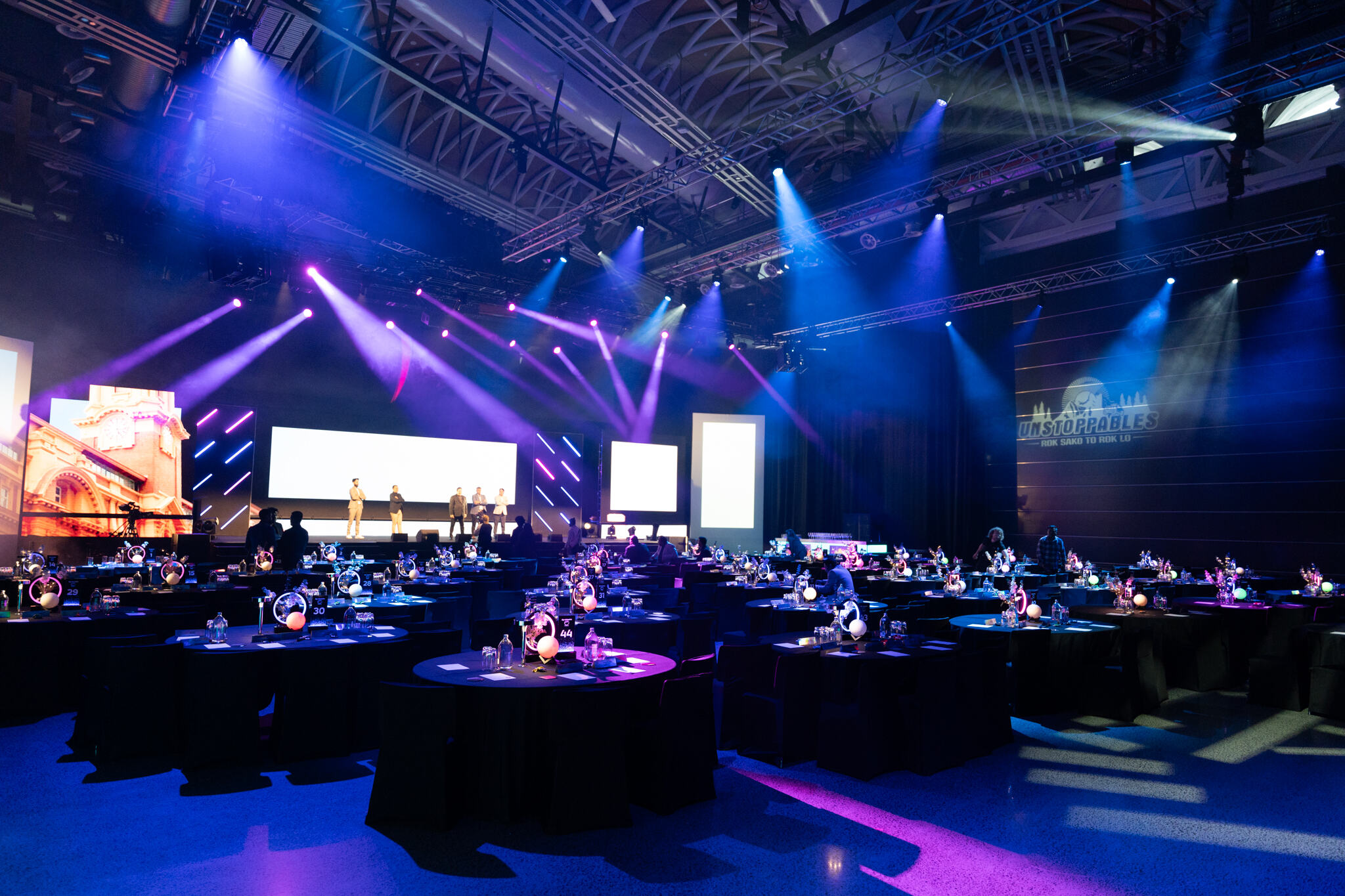 A large event hall with round tables and chairs is set up for a gala or award ceremony. Colorful stage lights illuminate a stage with people standing in front of large screens, and decorative lighting adds a festive atmosphere.