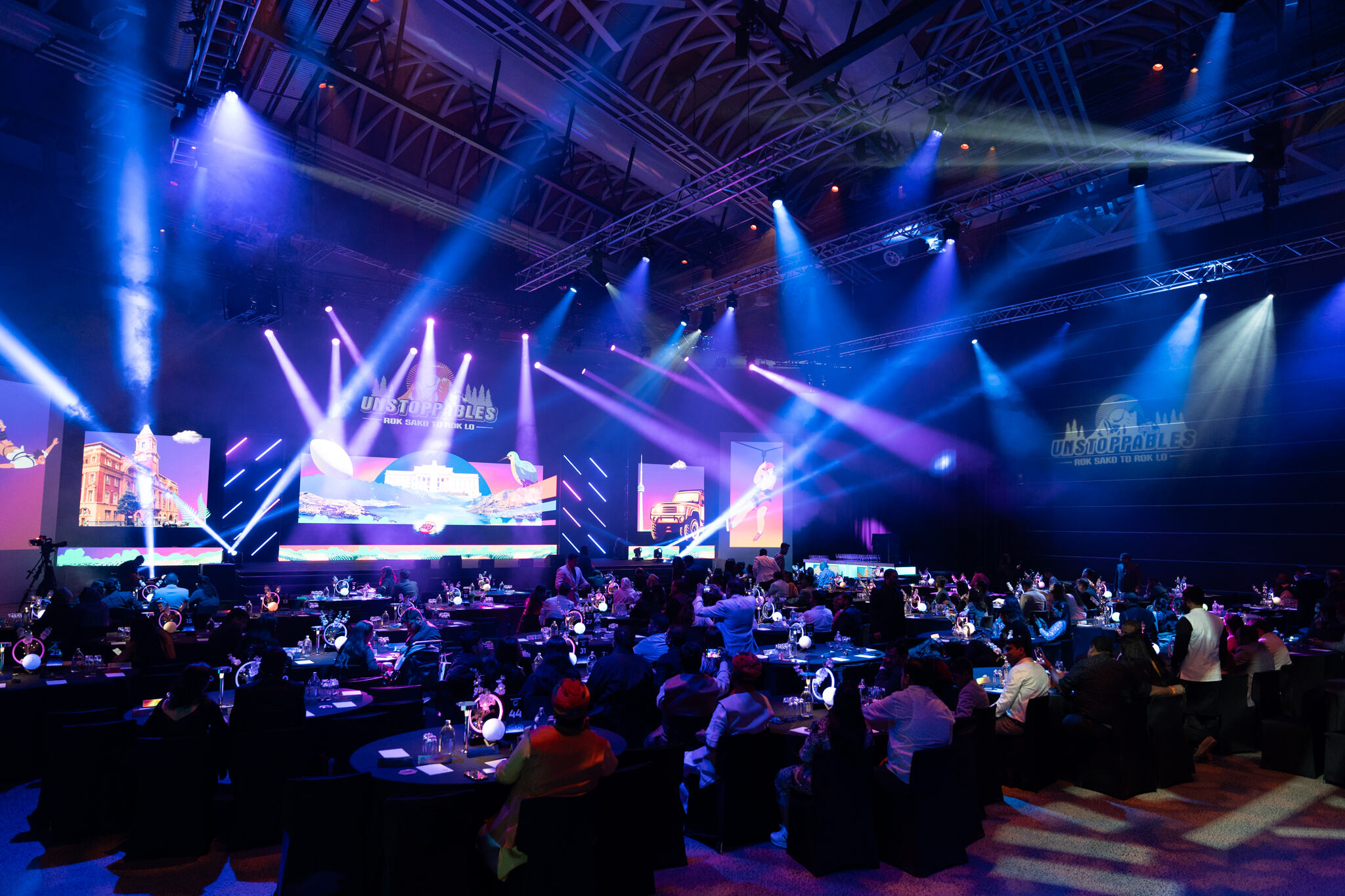 A large indoor event with round tables and seated guests faces a brightly lit stage with colorful lights, large screens, and the word UNSTOPPABLES projected on the wall.