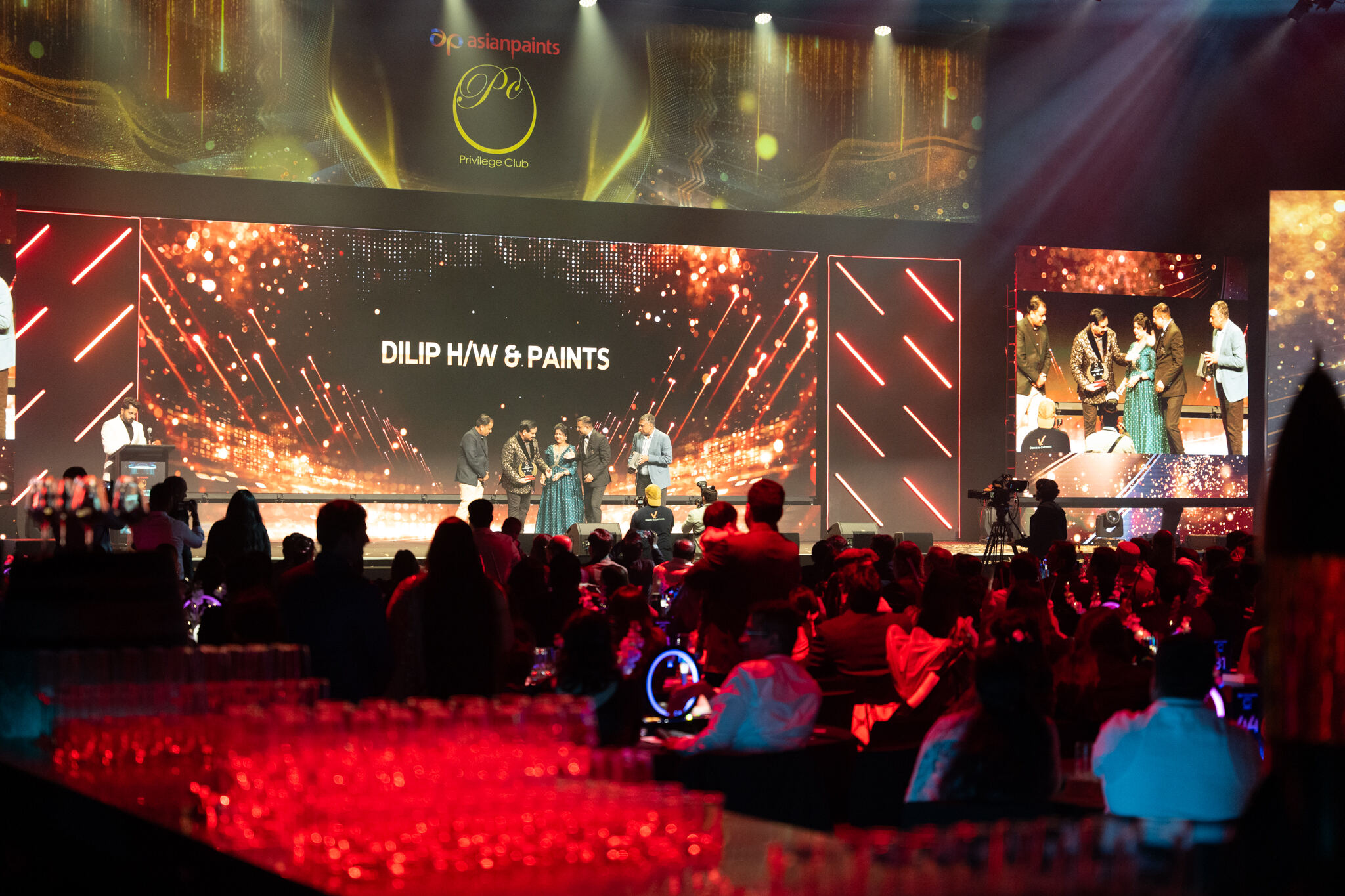 A large audience watches an awards ceremony on stage, where several people stand under a screen displaying DILIP H/W & PAINTS. The stage is brightly lit with colorful lights and event branding.