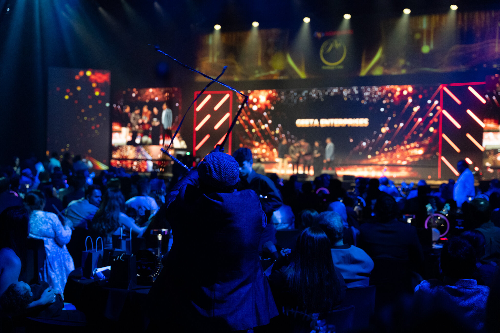 A crowd of people in a dimly lit event hall watches a brightly lit stage where an award ceremony is taking place. Large screens display GETTA ENTERPRISES and colorful lights illuminate the venue.