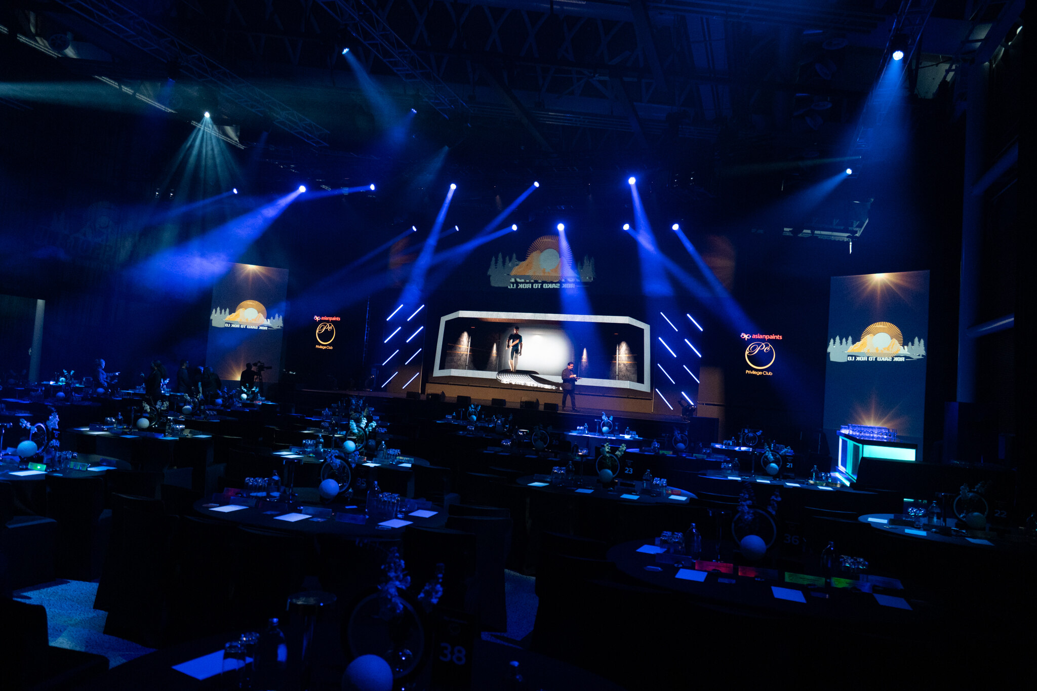 A dark, elegant event hall with blue stage lighting, round tables set for guests, and a large stage featuring screens displaying a sun logo and presenters standing in the spotlight.