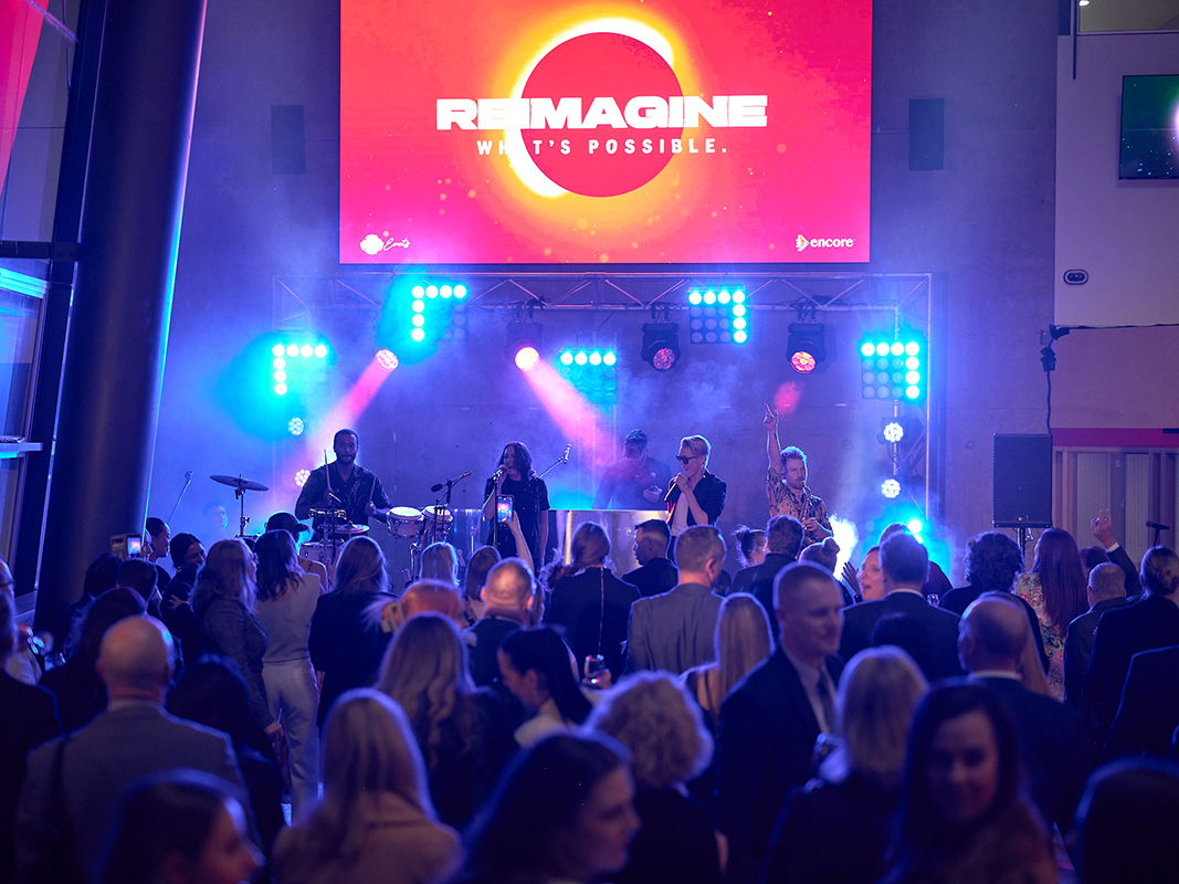 A crowd watches a live band perform on stage under colorful lights. A large screen above the stage displays the words REIMAGINE WHATS POSSIBLE. People in formal attire fill the foreground.