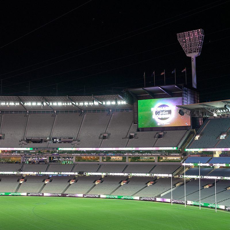 A wide view of an empty stadium at night, with rows of gray seats, a large green field, and a lit scoreboard displaying graphics—perfect for an event company Melbourne or AV company Melbourne to host spectacular live productions.