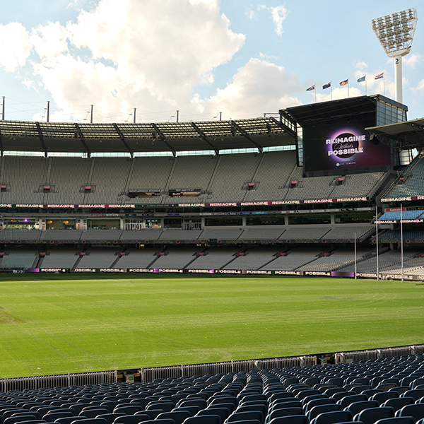 A large, empty sports stadium with rows of seats, a green field, and a big screen displaying REIMAGINE POSSIBLE. Flags fly atop the structure under a partly cloudy sky.
