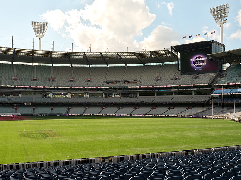 A large, empty sports stadium with rows of blue seats, a green field, and a big screen displaying the words IMAGINE THE POSSIBLE under a partly cloudy sky.