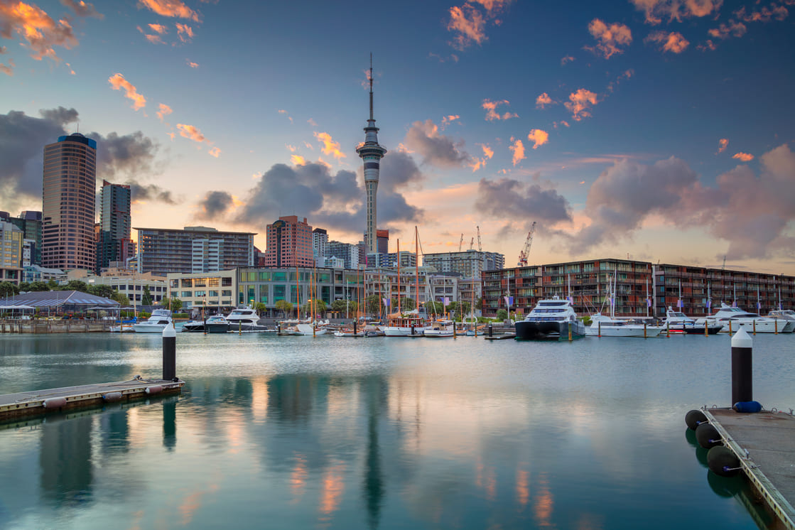 A harbor with docked boats reflects city buildings and the tall Sky Tower at sunset, with colorful clouds in the sky and calm water in the foreground.