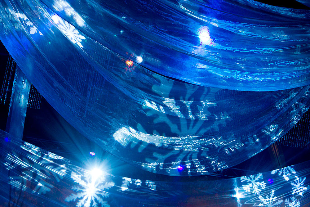 Blue translucent fabric draped from the ceiling with projected white snowflake patterns and bright blue lights, creating a wintry, festive atmosphere.