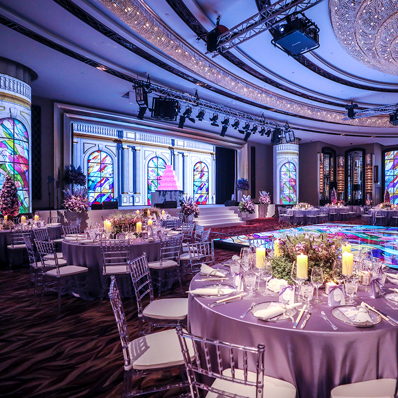 A lavishly decorated banquet hall set for weddings, with round tables adorned with candles and elegant tableware, a colorful illuminated dance floor, and a stage with stained glass window projections and a pink cake centerpiece.